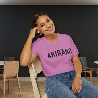 Person wearing a pink 'ARIRANG' t-shirt sitting in a room with chairs and tables. - Bangtan Tour 2026 Unofficial Shirt [Bias Arirang Color Vinyl Edition, Simple Text] - Jung Kook, JK, Orchid