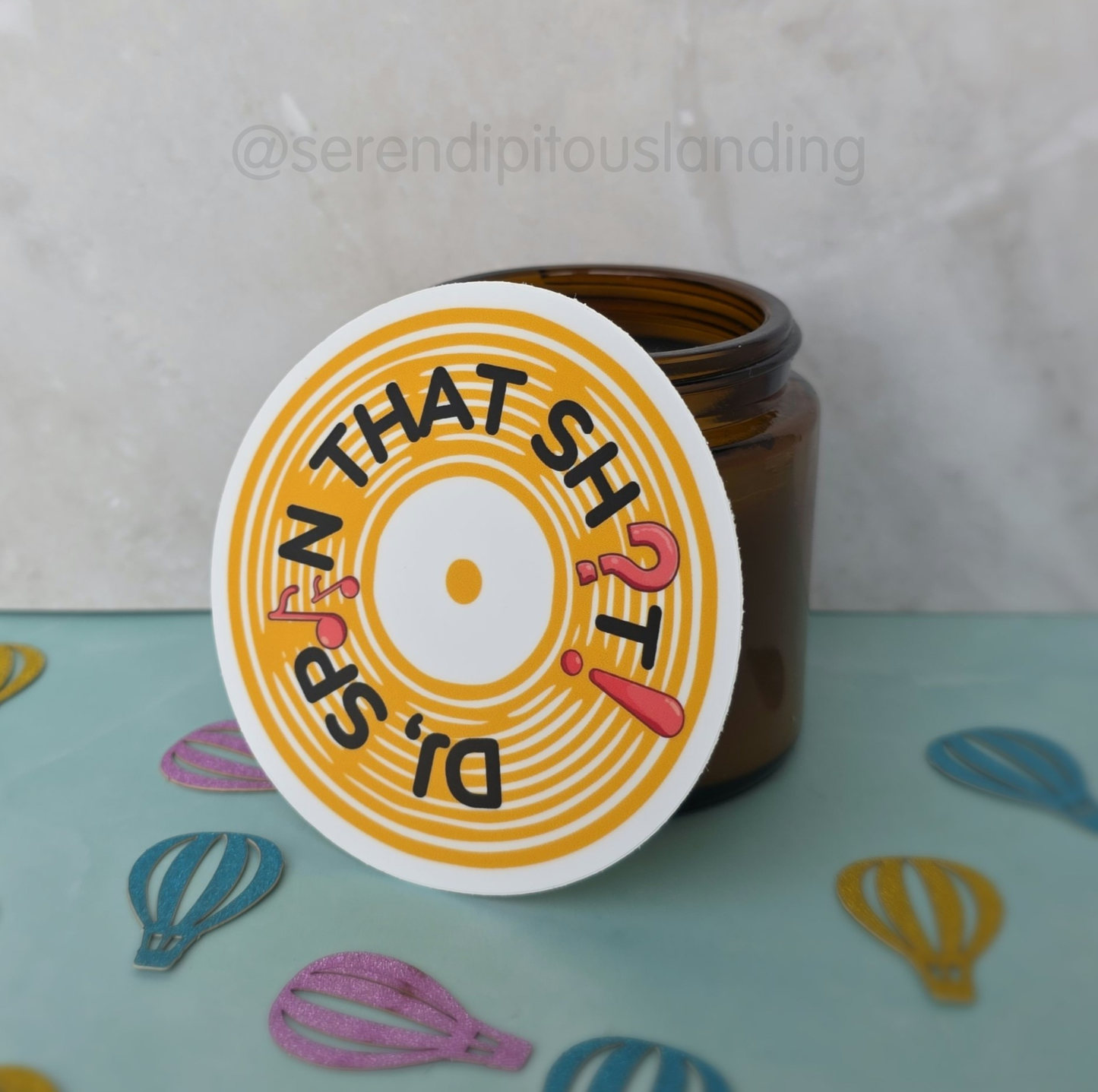 Brown jar with a colorful label on a light blue surface with decorative elements. - DJ Spin That Sticker | Serendipitous Landing
